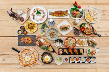 Ahşap masa üzerinde Uluslararası Gıdalar üst görünümü Suşi, Sashimi, Nachos, Spagetti, Naan, Sandviç, Erişte, Biftek, Ramen içerir. Yakitori, Pizza, Bibimbap, Kızarmış Tavuk Kanatları, Brownie Frappe.
