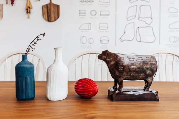 Wooden cow sculpture with cut of meat beef of diagram on it with blue, white jar and red fruit on wooden table.