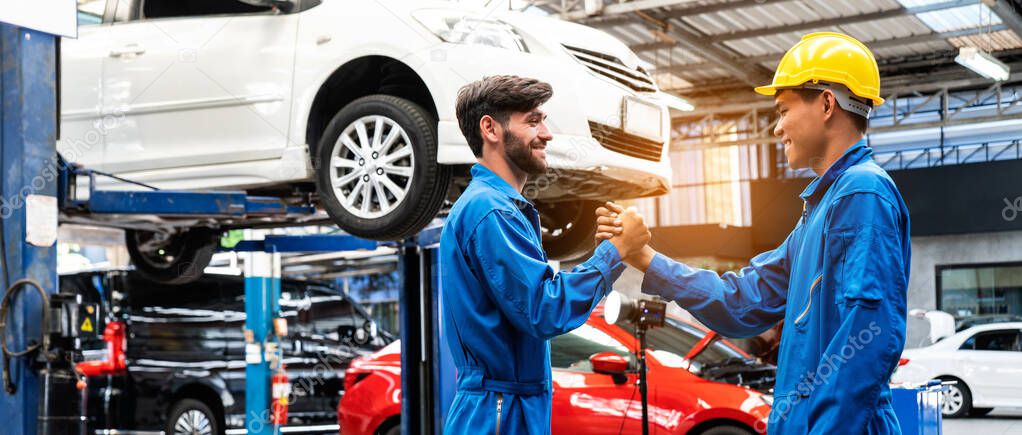 Mechanic in blue work wear uniform, shake hands with his assistant with a lifted car in the background. Automobile repairing service, Professional occupation teamwork. Vehicle maintenance.
