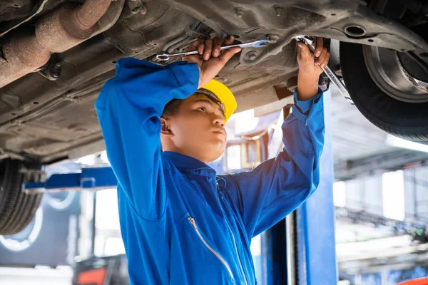 Asian mechanic wearing blue workwear and safety helmet examining the car bottom with wrenches. Auto car repair service center. Professional service.