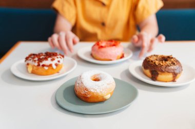 Masadaki çörekler arasında çilek pembesi, çikolata, şeker kaplı ve pastırma, masada peynir çöreği ve modern kafede arka planda sarı gömlek giyen kadın yer alıyor. Kadın yaşam tarzının keyfi.