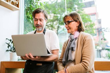 Önlüklü yetişkin bir adam, aydınlık pencerenin yanındaki yaşlı bir kadına dizüstü bilgisayarı gösterir, rahat bir kafede ya da dükkanda küçük bir iş danışmanlığı, eğitim ya da müşteri hizmetleri önerir..