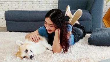 Genç bayan evcil hayvan sahibi yumuşak halıya uzanmış, gülümsüyor, kanepede gevşemiş Corgi yavrusunu okşuyor. Sıcak bir ev sahnesi, sıcak ve huzurlu bir oturma odasında köpek eşiyle insan arasındaki bağı gösteriyor. 4k video