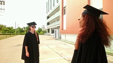 Şapka ve cüppeli iki mezun modern kampüs binasının önünde sarılıp gülümsüyor. Mutlu an, mezuniyet törenini, üniversite başarısını, üniversite başarısını, eğitim diplomasını yakalar. 4k video