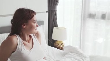 Parlak bir pencerenin yanında uzanan gülümseyen kadın, rahat bir yatak odasını nötr bir dekor ve komodinle dolduran, güne yeni bir başlangıç yakalayan ve rahatlayan ev konforu..