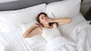 Beyaz atletli bir kadın beyaz çarşaflar ve yastıklarla yatakta uzanıyor, kolları açık, dinleniyor ve sabahları en az yatak odasında dinleniyor. 4k video
