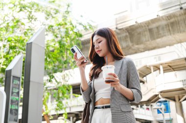Genç kadın, şehir merkezinin yakınındaki bir caddede buzlu kahve yudumlarken akıllı telefonu kontrol ediyor, modern şehir yaşam tarzını ve mobil bağlantılarını yakalıyor..
