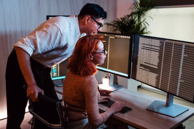 İki meslektaş, modern bir ofiste çift izleme masasında birlikte çalışıyor, geceleri kod veya verileri gözden geçiriyorlar. Biri durur ve yol gösterir, diğeri ise düşük ışıkta parlayan ekranlar..