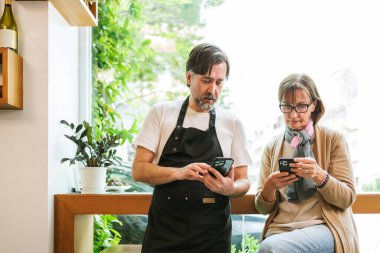 İki yetişkin küçük işletme sahibi, pencerenin kenarında rahat bir kafede akıllı telefonlarını birlikte kullanırlar; biri önlük giyer. Doğal ışık ve bitkiler gelişigüzel iş yerini çerçeveliyorlar..