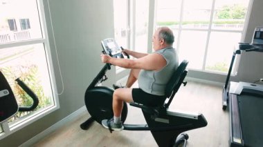 Kıdemli bireysel egzersizler parlak spor salonunda yatay duran bisiklet üzerinde. Sahne refahı, gücü, duruşu, hareketliliği, uygunluğu, performansı ve refahı artırmak için sağlık denetimini gösteriyor..