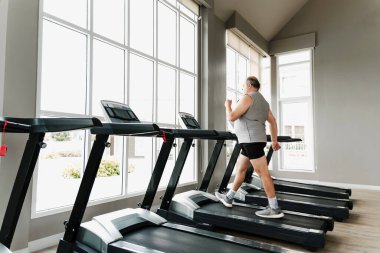 Adam büyük pencereli, ferah spor salonunda koşu bandında yürüyor. Sahne, kardiyo fitness, sağlık, refah, motivasyon, performans, koçluk, güç, koşu, koşu ve rehabilitasyon gösteriyor..