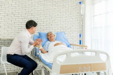 Adam hastane yatağında gülümseyen yaşlı bir hastayla el ele tutuşup rahatlık ve destek teklif ediyor. Parlak oda güven, iyileşme, iyileşme, umut, hemşire bakımı, yardım, refah ve birlikteliği yansıtır.