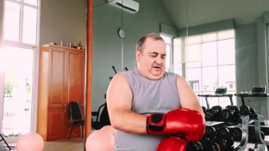 Adam spor salonundaki bankta spor kıyafetleriyle çevrili boks eldivenlerini çıkarıyor. Parlak alan iyileşmeyi, koçluğu, gücü, terlemeyi, çevikliği, disiplini, uygulamayı, esnemeyi ve sağlığa odaklanmayı yansıtır. 4k