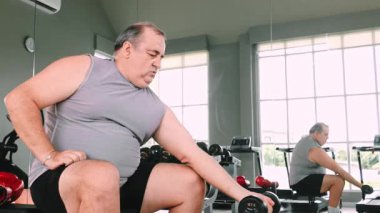 Yetişkin bir adam spor salonunda bir biseps kıvrımında halterini kaldırıyor, bir duvar aynası önünde bankta oturuyor, kararlılık, fitness eğitimi ve kapalı bir çalışma ortamında güç arttırmayı vurguluyor..