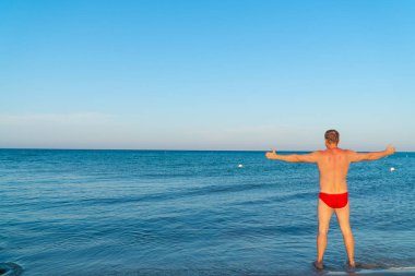 Sahilde kırmızı mayo yaz genç ve yakışıklı kaslı adam, denize bakarak, Akdeniz'de başparmak ile kol yanlara yayıldı