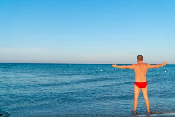 Young and handsome  muscular man in red swimsuit summer on the beach, looking at the sea, Spread arm sideways with thumbs up in Mediterranean