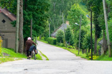 Yaz aylarında küçük bir kasaba yolu boyunca bisiklet iterek yaşlı bir kadının arka görünümü
