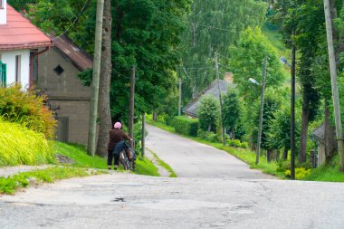 Yaz aylarında küçük bir kasaba yolu boyunca bisiklet iterek yaşlı bir kadının arka görünümü