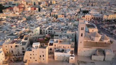 Eski Katedral San Corrado (Duomo Vecchio) ile Molfetta rıhtımında uçan İHA 'dan güzel panoramik 4K video, gemi ve yatlı liman ve Molfetta şehir günbatımı. Apulia, İtalya