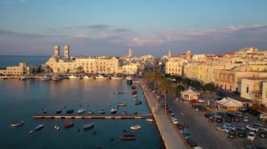 Eski Katedral San Corrado (Duomo Vecchio) ile Molfetta rıhtımında uçan İHA 'dan güzel panoramik 4K video, gemi ve yatlı liman ve Molfetta şehir günbatımı. Apulia, İtalya