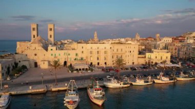 Eski Katedral San Corrado (Duomo Vecchio) ile Molfetta rıhtımında uçan İHA 'dan güzel panoramik 4K video, gemi ve yatlı liman ve Molfetta şehir günbatımı. Apulia, İtalya