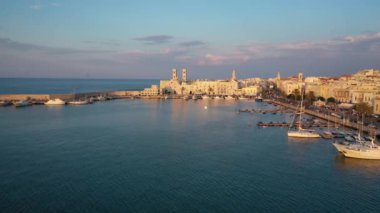 Eski Katedral San Corrado (Duomo Vecchio) ile Molfetta rıhtımında uçan İHA 'dan güzel panoramik 4K video, gemi ve yatlı liman ve Molfetta şehir günbatımı. Apulia, İtalya