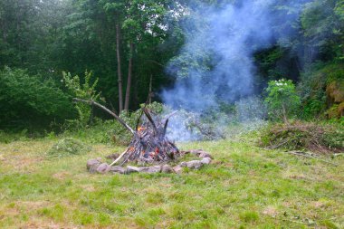 Kamp alanında duman bombası. Etrafı taşlar, yeşil çimenler ve ağaçlarla çevrili bir şömine..