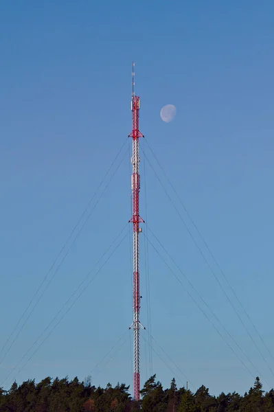 High red and white cellular tower at daytime with moon shining next to the top of the tower.