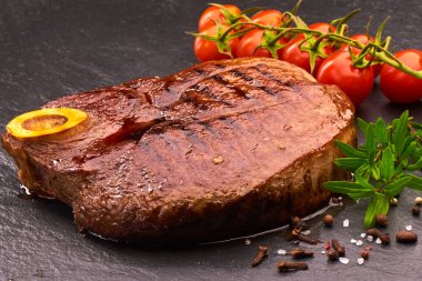 Siyah taş tahtada ızgara biftek, kiraz domatesleri ve baharatlar.