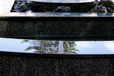 Katmanlı taştan bir fıskiyenin sakin bir yakın çekimi, su yavaşça çağlayarak akıyor ve etrafındaki yeşilliği ve gökyüzünü yansıtıyor. Sahne, bahçedeki huzur ve doğal güzelliği çağrıştırıyor..