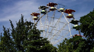 Eski bir dönme dolap, parlak mavi bir gökyüzüne karşı dimdik durur. Renkli kamaraları eski bir çağı işaret eder. Ağaçlar yolculuğu çerçeveler, nostaljik ve biraz melankolik atmosfere ekler..