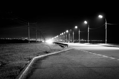 Karanlık bir gökyüzünün altındaki uzak bir şehrin siluetine doğru yol alan sokak lambalarıyla dolu uzun bir yolun siyah ve beyaz gece çekimi. Görüntü karamsar ve sinematiktir..