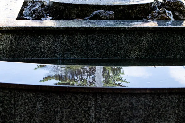 Katmanlı taştan bir fıskiyenin sakin bir yakın çekimi, su yavaşça çağlayarak akıyor ve etrafındaki yeşilliği ve gökyüzünü yansıtıyor. Sahne, bahçedeki huzur ve doğal güzelliği çağrıştırıyor..