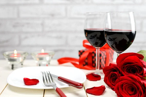 Sevgililer Günü yemeği konsepti. İki kadeh şarap, kırmızı güller, hediye kutusu, tabak ve yanan mumlar beyaz ahşap bir masada. Seçici odak.