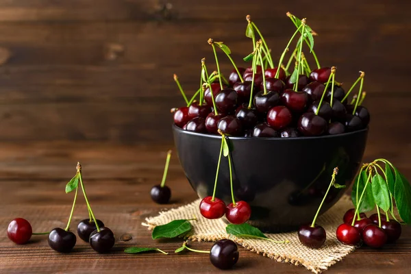 Fresh sour cherries in black bowl and green leaves on brown wooden table  with copy space for text. Fresh ripe sour cherries.