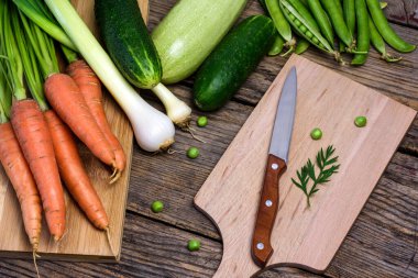 Çeşitli taze sebzeler. Bezelyeler, havuçlar, salatalık, kabak ve soğanlı eski ahşap bir masada tahta tahta ve mutfak bıçağı..