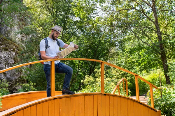 young bearded millennial guy a traveler in a hat with a backpack stands on a wooden bridge, studies the route and looks for the road on the map, got lost