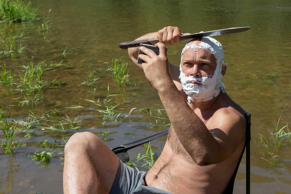 crazy or mad man shaves his head with large long knives, machetes. outdoors, camping sitting on a chair