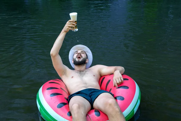 bearded young guy, hipster catches beer foam in his mouth, swims in a rubber ring. enjoys relaxing, resting in nature.