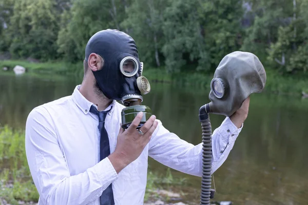 man in a shirt and tie, in a gas mask holds in his hand a second gas mask. Sorry about a friend, says goodbye. Outdoors on the background of the river and the forest