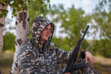 kamuflaj bir silahla tek tip adam çölde bir ağacın altında oturuyor.