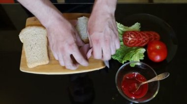 Genç adam kendini iki parça ekmek, sosis, yeşil salata ve domates lezzetli ve sulu sandviç yapar ve bir üçgen şeklinde iki bölüme keser, siyah bir cam masa arka plana karşı, yakın çekim
