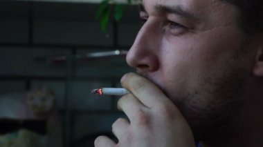 Yüzünde bir inlemek ile genç bir yakışıklı beyaz adam balkonda bir sigara içiyor ve duman, yakın çekim, yavaş hareket havaya uçurmak