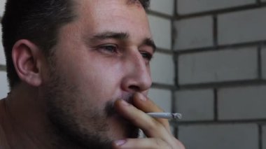Yüzünde bir inlemek ile genç bir yakışıklı beyaz adam balkonda bir sigara içiyor ve duman, yakın çekim, yavaş hareket havaya uçurmak
