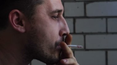 Yüzünde bir inlemek ile genç bir yakışıklı beyaz adam balkonda bir sigara içiyor ve duman, yakın çekim, yavaş hareket havaya uçurmak
