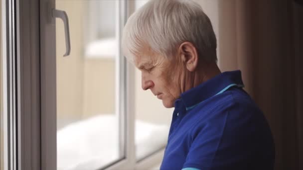 Lonely senior man standing near the window while wearing glasses and looking outdoors — Vídeo de stock