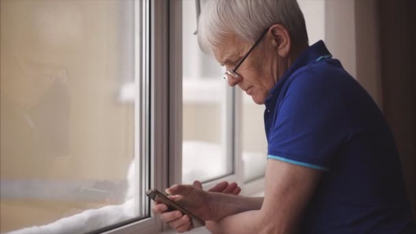 Senior concentrated man in glasses writing message on his brand-new smartphone staying near window at home. Indoors. — Vídeos de Stock