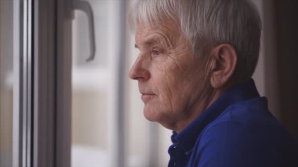 Позитивний пенсіонер дивиться на вікно, стоячи біля вікна вдома. Він у сучасному блакитному поло. — стокове відео