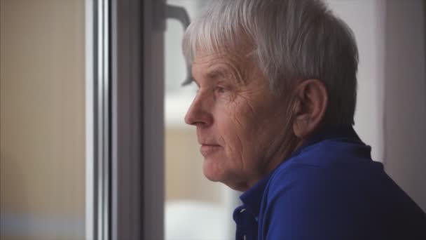 青の目で高齢者の灰色の男の顔にクローズアップ肖像画。彼は窓の近くを見て何かを考えて — ストック動画