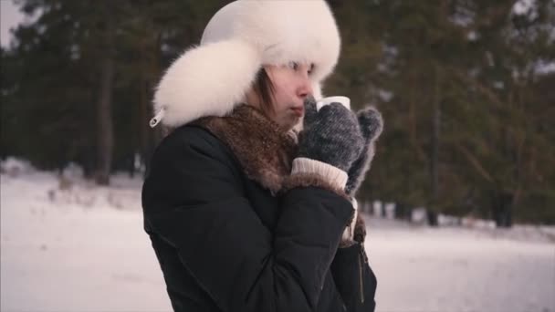 Young model in winter white hat enjoys the snow and drinks hot beverage standing outside forest. Girl enjoying winter outdoors — Stock video